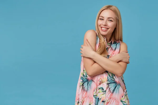 Positivo bonito jovem senhora de cabelos longos com penteado casual abraçando-se suavemente e sorrindo alegremente para a câmera, posando sobre fundo azul em vestido romântico florido — Fotografia de Stock