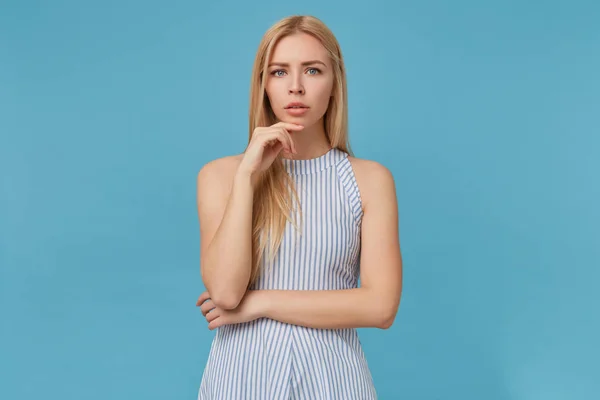 Foto interior de grave jovem de cabelos compridos fêmea em vestido de verão romântico inclinando-se queixo na mão levantada, isolado sobre fundo azul, olhando para a câmera com rosto grave — Fotografia de Stock