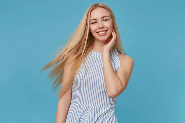 Retrato de jovem senhora loira atraente com cabelos longos mantendo a mão em seu rosto e sorrindo alegremente para a câmera, posando sobre fundo azul, demonstrando seus dentes brancos perfeitos — Fotografia de Stock