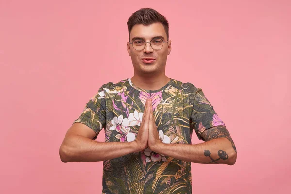 Όμορφος νεαρός άνδρας φορώντας γυαλιά και άνθος t-shirt στέκεται με διπλωμένες παλάμες πάνω από ροζ φόντο, κάνοντας namaste χειρονομία, αναζητούν ηρεμία και ικανοποίηση — Φωτογραφία Αρχείου