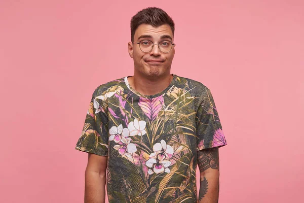Απογοητευμένος νεαρός άνδρας με κοντά μαλλιά φορώντας γυαλιά και άνθος t-shirt, στέκεται πάνω από ροζ φόντο με τα χέρια κάτω, συστέλλεται μέτωπο και pursing χείλη — Φωτογραφία Αρχείου