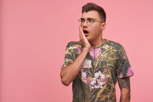 Close-up shot of shocked short haired handsome guy looking aside with jaw dropped out like seeing something unexpected, holding palm on his cheek, standing over pink background — Stock Photo, Image