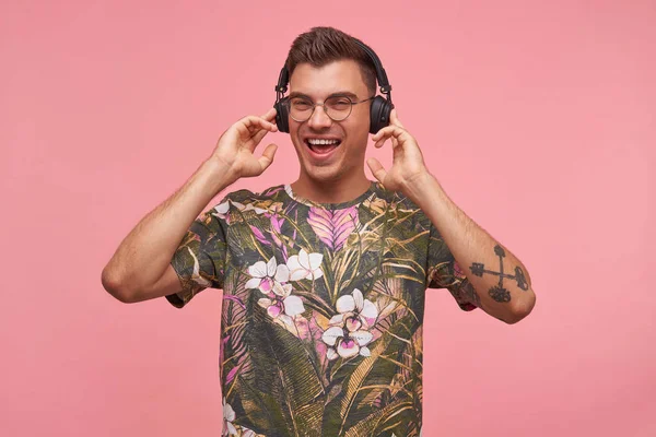 Εσωτερική λήψη του όμορφου νεαρού άνδρα φορώντας γυαλιά και άνθος t-shirt στέκεται πάνω από ροζ φόντο με υψωμένα τα αυτιά χέρια, απολαμβάνοντας μουσική στα ακουστικά του, αναζητούν ευτυχισμένη και ευχάριστη — Φωτογραφία Αρχείου