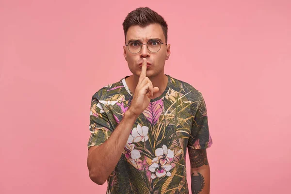 Κοντινό πλάνο του ελκυστική νεαρός άνδρας με τατουάζ κρατώντας δείκτη στα χείλη, ζητώντας να κρατήσει μυστικό, δεν κάνουν θόρυβο, επιδεικνύοντας χειρονομία σιωπής, στέκεται πάνω από ροζ φόντο — Φωτογραφία Αρχείου
