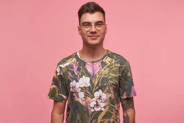 Εσωτερικό πορτραίτο ενός όμορφου νεαρού με τατουάζ που φοράει γυαλιά και λουλουδάτο μπλουζάκι, χαμογελώντας απαλά στην κάμερα, απομονωμένο σε ροζ φόντο — Φωτογραφία Αρχείου