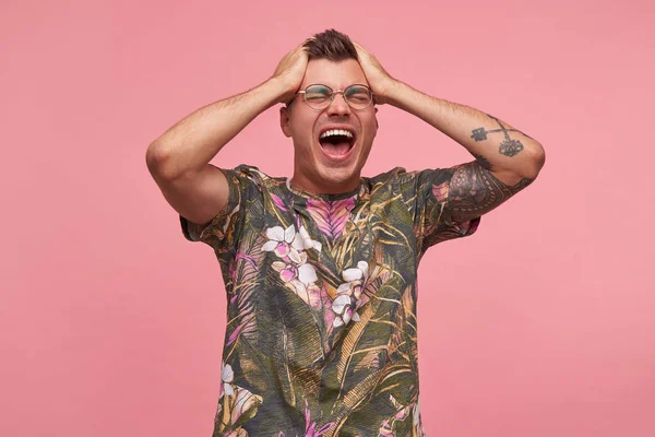 Excited attractive guy with tattoos in casual t-shirt posing over pink background, standing with palms on his head, closing eyes and shouting loud — Stock Photo, Image