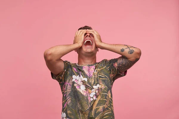 Studio shot of flurried handsome guy standing with thrown back head, closing eyes with his palms and opening mouth widely, isolated over pink background