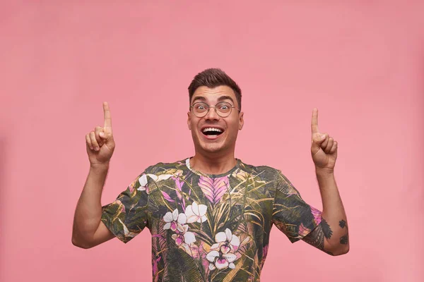 Studio φωτογραφία του γοητευτικού νεαρού κοντό μαλλιά αρσενικό σε ανθισμένο t-shirt κοιτάζοντας στην κάμερα χαρούμενα, δείχνοντας με τα δάχτυλα προς τα πάνω, απομονώνονται σε ροζ φόντο — Φωτογραφία Αρχείου