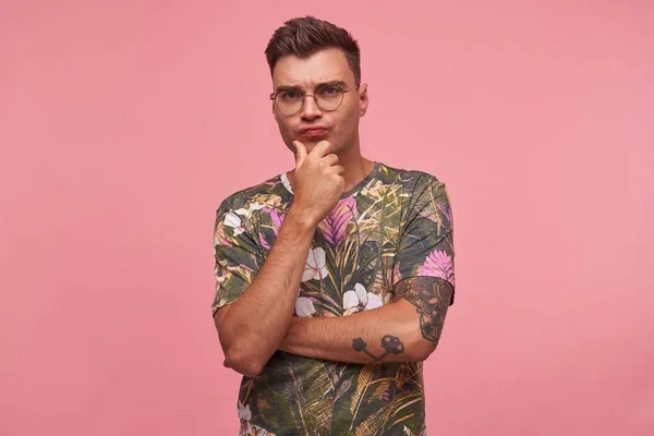 Πορτρέτο του σκεπτικού όμορφος νεαρός άνδρας σε ανθισμένο t-shirt με σοβαρό πρόσωπο, κρατώντας το πηγούνι του με το χέρι και συνοφρύωμα στην κάμερα πάνω από ροζ φόντο — Φωτογραφία Αρχείου