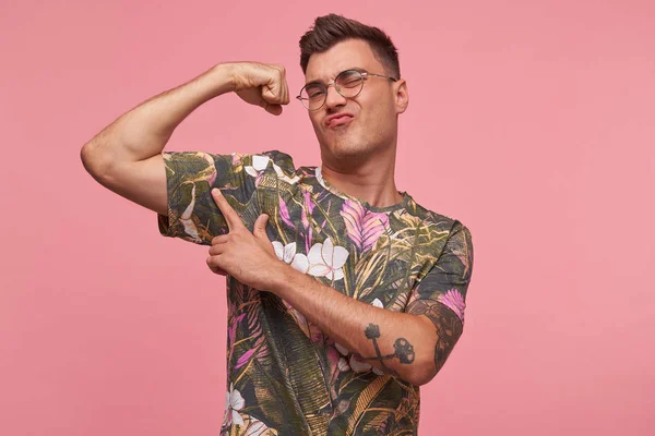 Studio shot of proud handsome tattooed young man in colored casual t-shirt pointing and showing his strong bicep, isolated on pink background — Stock Photo, Image