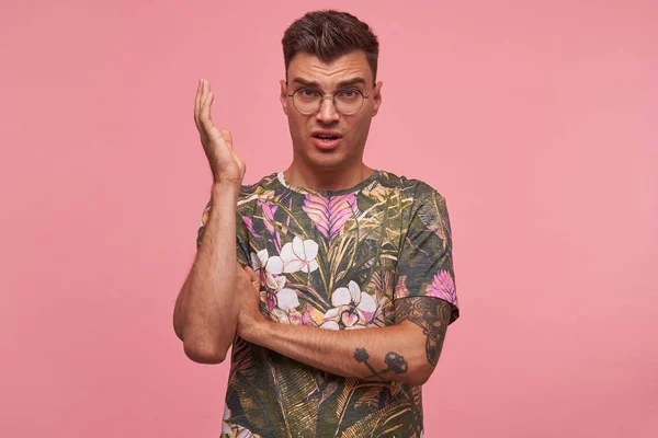 Ελκυστικός νεαρός άνδρας με μοντέρνο κούρεμα φορώντας γυαλιά και άνθος t-shirt σηκώνει το χέρι του και κάνει αγανακτισμένος πρόσωπο, συνοφρυωμένος εγκάρσια, απομονωμένος σε ροζ φόντο — Φωτογραφία Αρχείου