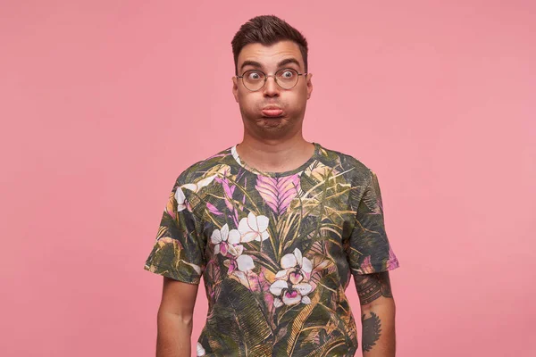 Εσωτερική πορτρέτο του αστεία νεαρός όμορφος tatooed αρσενικό με γυαλιά grimacing και κάνοντας πρόσωπα, θέτοντας πάνω από ροζ φόντο με τα χέρια κάτω — Φωτογραφία Αρχείου
