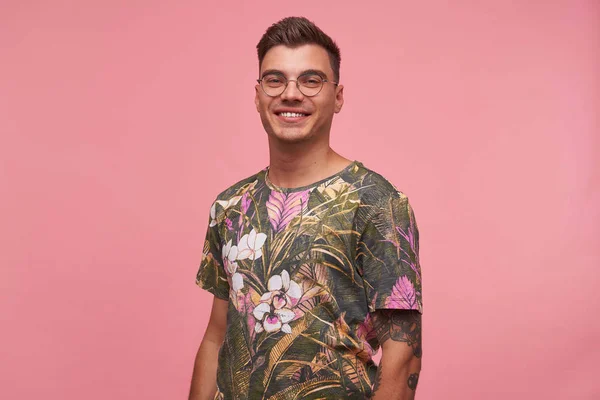 Στούντιο πλάνο του όμορφου νεαρού άνδρα με τατουάζ με γοητευτικό χαμόγελο ποζάροντας πάνω από ροζ φόντο, κοιτάζοντας με χαρά στην κάμερα, φορώντας γυαλιά και λουλουδάτο t-shirt — Φωτογραφία Αρχείου