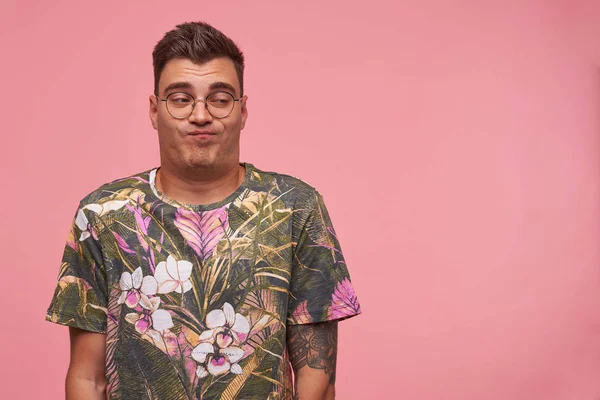 Αστεία όμορφη νεαρός άνδρας σε λουλουδάτο t-shirt και γυαλιά κοιτάζοντας κατά μέρος αδιάφορα, πτυσσόμενα χείλη και κάνοντας απαθές πρόσωπο, στέκεται πάνω από ροζ φόντο — Φωτογραφία Αρχείου