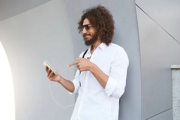 Beau jeune homme joyeux bouclé debout sur un mur gris avec une tablette à la main, ayant un appel vidéo avec écouteurs, portant des lunettes de soleil et des vêtements décontractés — Photo