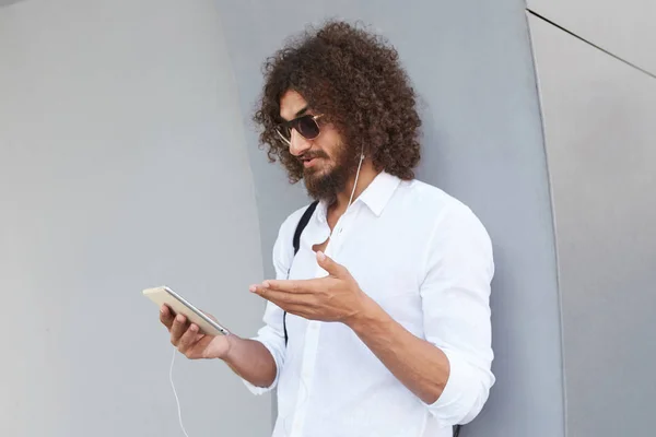 Attrayant jeune barbu cheveux foncés bouclés mâle appuyé sur le mur extérieur gris pendant le chat vidéo avec tablette, portant des lunettes de soleil et des vêtements décontractés — Photo