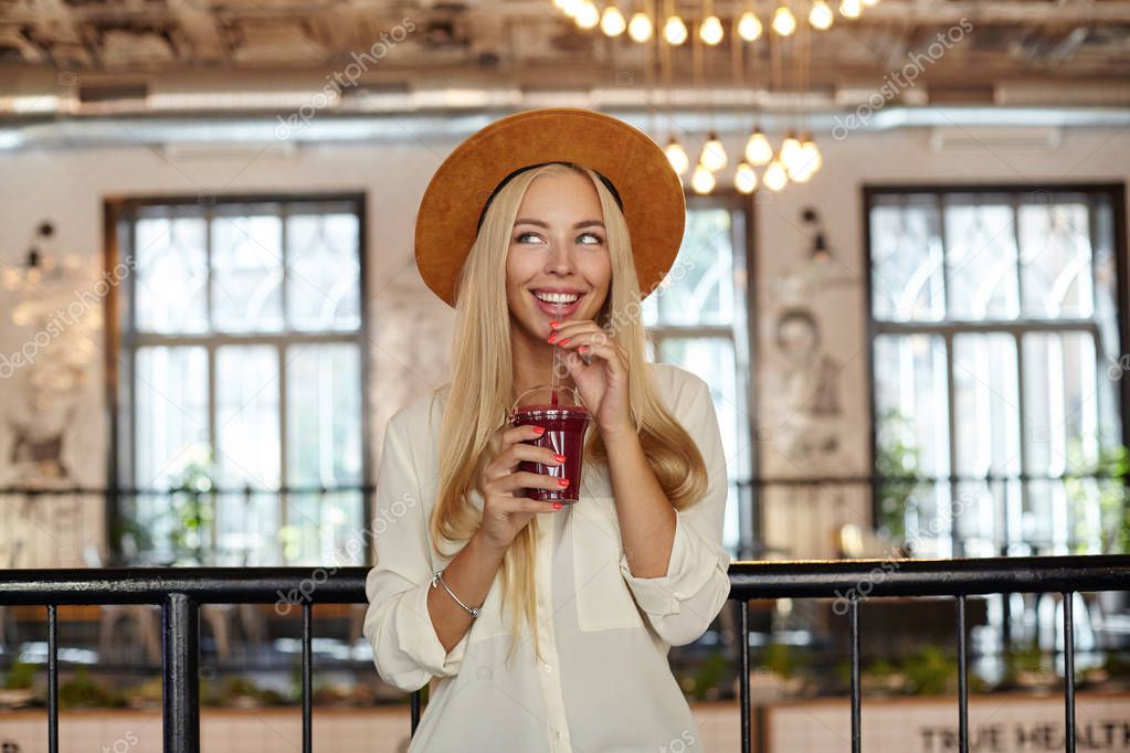 Indoor shot of happy young blonde woman with blue eyes standing over restaurant interior, drinking lemonade with straw and looking aside with dreamy smile