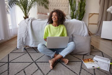 Cheerful young dark skinned woman sitting on floor in casual clothes, studying with texbooks and modern laptop, smiling joyfully and being in nice mood