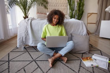 Indoor photo of beautiful young dark skinned woman with brown curly hair sitting on carpet in bedroom with laptop on her legs, wearing casual clothes, positive mood