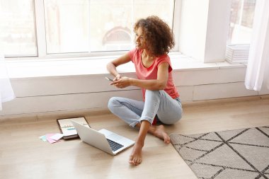 Güzel, koyu tenli, kahverengi kıvırcık saçlı, bacak bacak üstüne atmış, pencere kenarına yaslanmış ve akıllı telefon tutan, düşünceli bir şekilde ileriye bakan bir kadın.