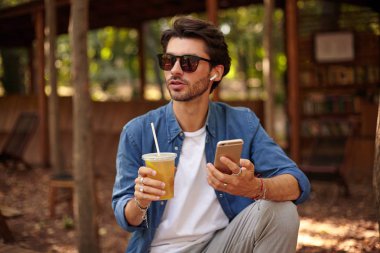 Sakallı genç, hoş bir erkek, öğle yemeği molası veriyor, elinde buzlu çayla şehir bahçesinde poz veriyor, cep telefonuyla arama yapıyor.