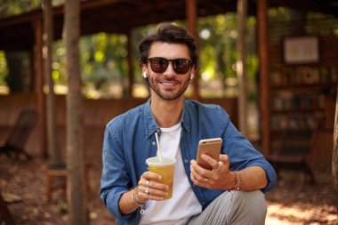 Güneş gözlüklü, halka açık bir yerde poz veren sevimli sakallı bir erkek portresi. Çekici bir gülümsemeyle kameraya bakıyor. Elinde cep telefonu ve elinde meyve suyu tutuyor.
