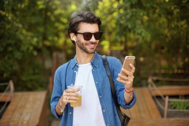 Yeşil şehir bahçesinde yürüyen, limonata içen, elinde cep telefonu tutan, ekranlara bakıp neşeyle gülümseyen sakallı genç bir erkek.