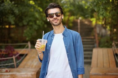 Güneşli sıcak bir günde yeşil şehir parkında poz veren rahat giyinmiş, elinde bir fincan limonata tutan ve kameraya gülümseyen güzel siyah saçlı bir adam.