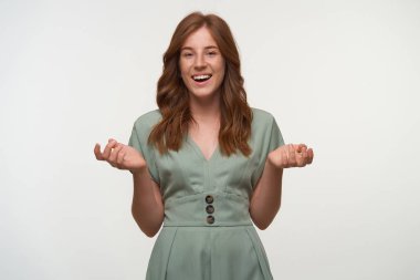 Mutlu kızıl saçlı güzel kadın pastel renkli klasik elbiseler giyiyor, kameraya neşeyle gülümsüyor ve avuçlarını yukarı kaldırıyor, beyaz arka planda soyutlanıyor.