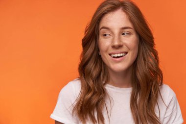 Beyaz tişörtlü, kıvırcık kırmızı saçlı, neşeli bir kadınla kapalı çekimde büyüleyici bir gülümsemeyle turuncu arka planda poz veriyor.