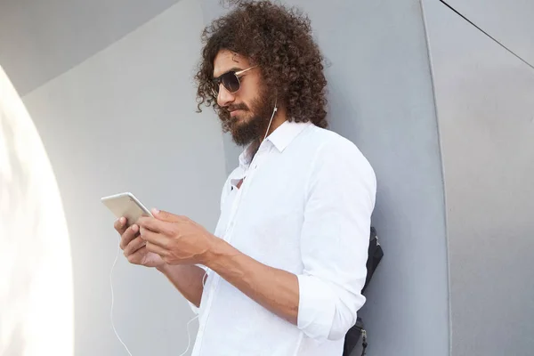 Portrait extérieur d'un homme bouclé attrayant debout au-dessus d'un mur gris, lisant des nouvelles sur une tablette en attendant quelqu'un, portant des vêtements et des lunettes décontractés — Photo