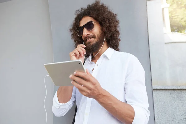 Plan en plein air avec beau jeune homme bouclé tenant la tablette à la main et ayant une conversation en ligne avec casque, portant des lunettes de soleil et chemise blanche — Photo