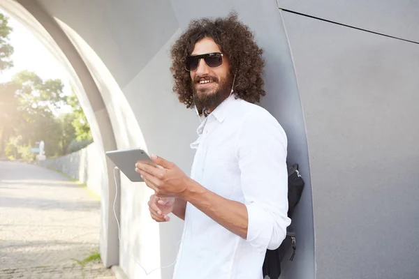 Jeune homme bouclé attrayant avec la barbe souriant doucement, écoutant de la musique avec des écouteurs sur sa tablette, posant sur un fond extérieur par une journée chaude ensoleillée — Photo