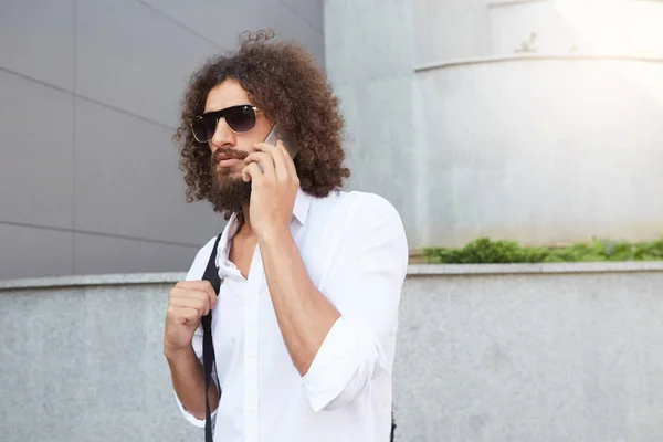 Serious good looking young curly man with beard walking down the street with smartphone in hand, wearing white shirt and black backpack