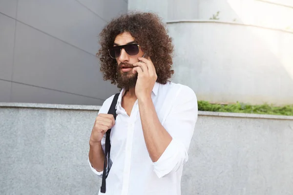 Beau jeune homme barbu aux cheveux bouclés foncés marchant dans la rue tout en ayant une conversation téléphonique importante, portant une chemise blanche et un sac à dos noir — Photo