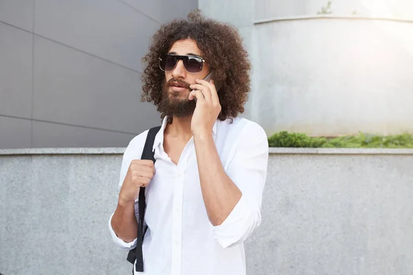Portrait extérieur d'un bel homme avec une barbe luxuriante et des boucles parlant au téléphone tout en marchant dans la rue, portant des vêtements décontractés — Photo