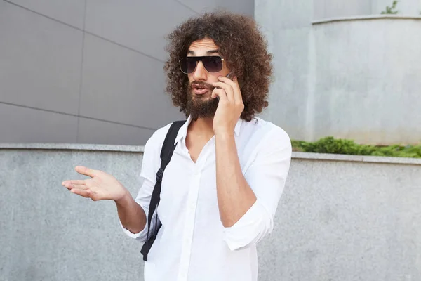 Jeune beau barbu aux cheveux bouclés posant sur le fond de la ville avec le visage agité, le front ridé et gesticulant tout en expliquant quelque chose au téléphone — Photo