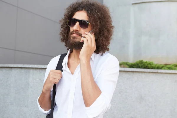 Outdoor shot of attractive young curly bearded male with phone in hand walking down the street on sunny day, wearing white shirt and black backpack