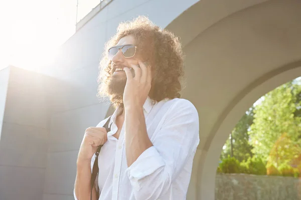 Beau jeune homme barbu ayant une conversation agréable au téléphone tout en marchant dans la rue verte par une journée chaude ensoleillée, portant des lunettes et une chemise blanche — Photo