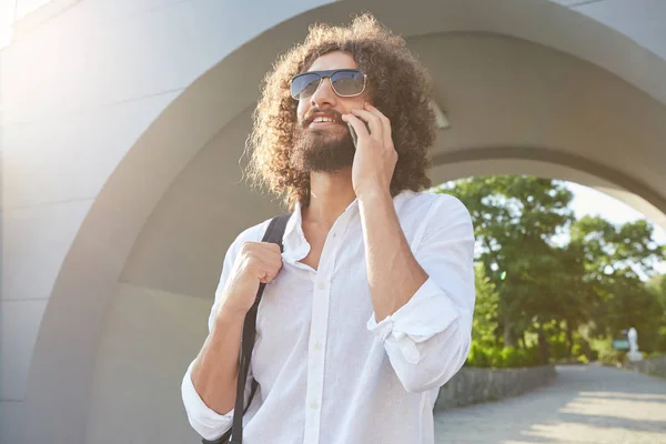Joyeux homme barbu aux cheveux bouclés portant des lunettes de soleil, marchant dans le parc de la ville le jour ensoleillé tout en parlant sur son téléphone portable, tenant son sac à dos — Photo
