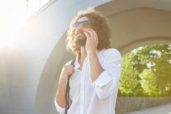 Plan extérieur d'un bel homme barbu avec des boucles parlant sur un téléphone portable, marchant dans le parc de la ville, portant des lunettes de soleil et des vêtements décontractés — Photo