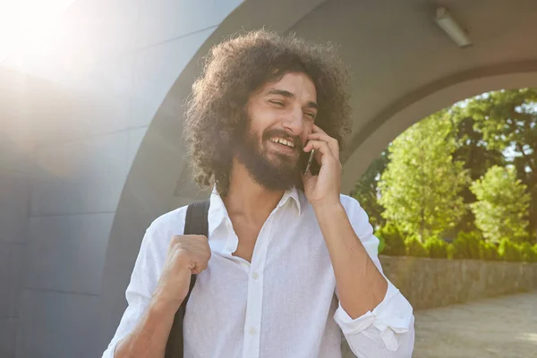 Jeune homme barbu attrayant aux cheveux bouclés bruns ayant une conversation agréable au téléphone, souriant joyeusement, posant sur le parc de la ville le jour ensoleillé — Photo