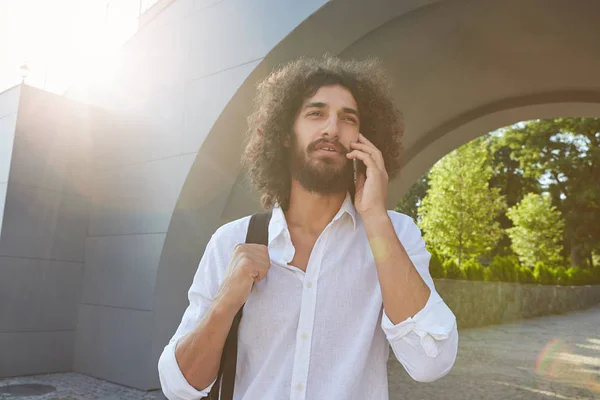 Jeune homme bouclé attrayant avec barbe marchant dans le parc de la ville, plissant du soleil et parlant au téléphone, portant une chemise blanche et un sac à dos noir — Photo