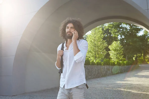 Portrait extérieur ensoleillé d'agréable jeune homme barbu marchant à travers l'arche dans le parc et parlant au téléphone, portant des vêtements décontractés et un sac à dos noir — Photo