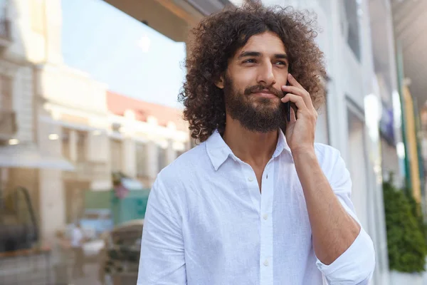 Portrait extérieur d'un beau jeune homme bouclé à la barbe luxuriante, parlant sur un téléphone portable pendant la pause déjeuner, portant une chemise blanche — Photo