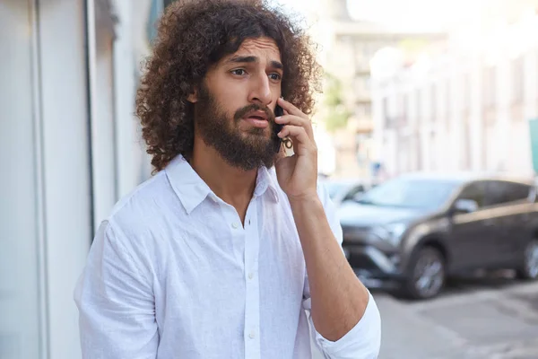 Portrait extérieur d'un homme barbu perplexe aux cheveux bouclés ayant une conversation téléphonique désagréable, debout sur le fond de la ville en chemise blanche — Photo