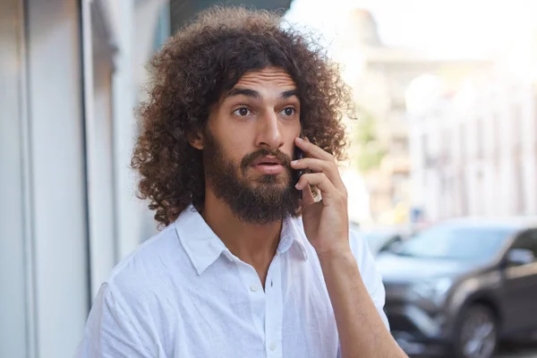 Surpris beau barbu aux cheveux bouclés bruns marchant dans la rue avec téléphone portable à la main, le front ridé et levant les sourcils — Photo