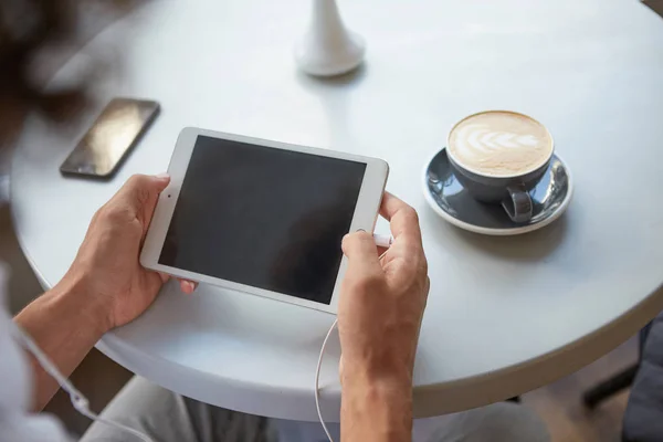 Primer plano interior de las manos del hombre sosteniendo la tableta con auriculares enchufados, almorzando con una taza de café, manteniendo el teléfono inteligente en la mesa — Foto de Stock