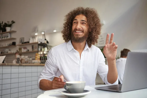 수염을 기르고 갈색 곱슬곱슬 한 머리를 하고 카페 탁자에 앉아 일하는 동안 커피를 마시며 친구와 인사하는 행복 한 젊은이의 실내 사진 — 스톡 사진