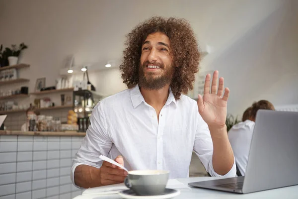 Cheerful lovely curly male with beard meeting familiar person and waving his hand, working remotely with laptop, posing over luncheonette interior